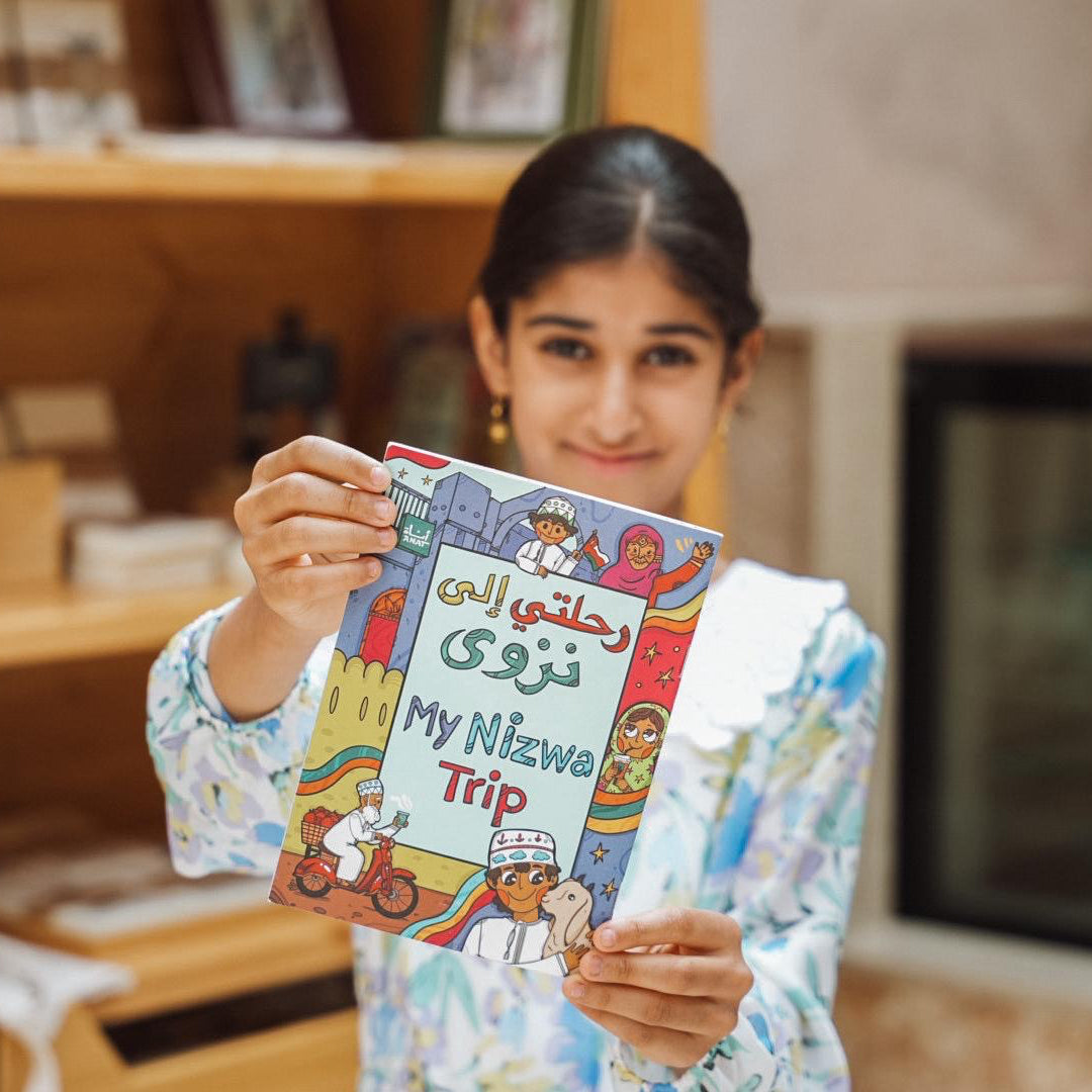 Girl holding a book titled 'My Nizwa Trip' in a home setting