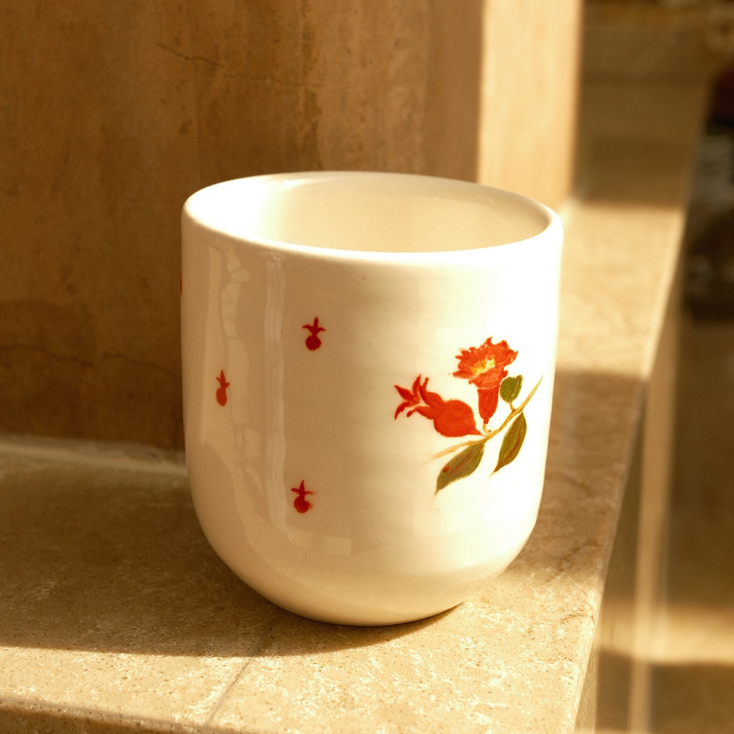 A white ceramic cup with a red floral and green leaf design, resting on a stone ledge in warm sunlight.