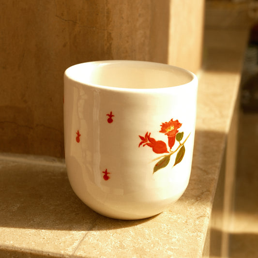 A white ceramic cup with a red floral and green leaf design, resting on a stone ledge in warm sunlight.