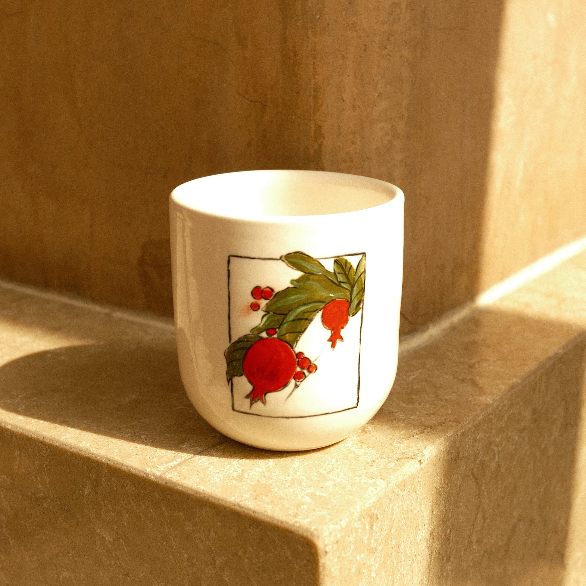 A white ceramic cup with a pomegranate illustration, resting on a beige stone ledge in warm sunlight.