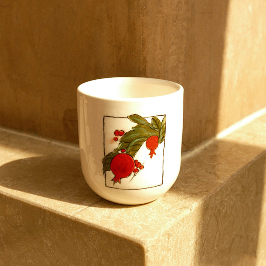 A white ceramic cup with a pomegranate illustration, resting on a beige stone ledge in warm sunlight.
