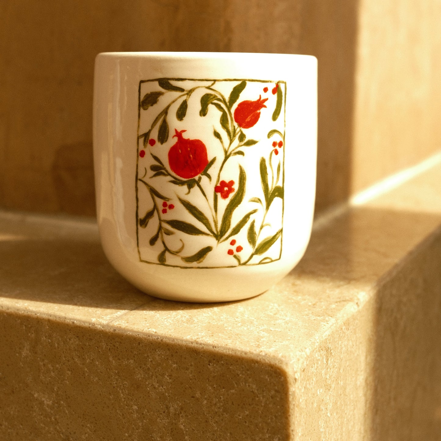 A white ceramic cup with a hand-painted pomegranate and leaf pattern, resting on a beige stone surface in warm sunlight.
