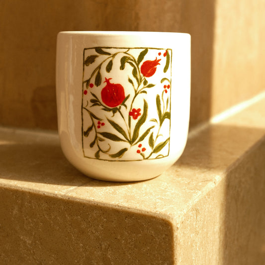 A white ceramic cup with a hand-painted pomegranate and leaf pattern, resting on a beige stone surface in warm sunlight.
