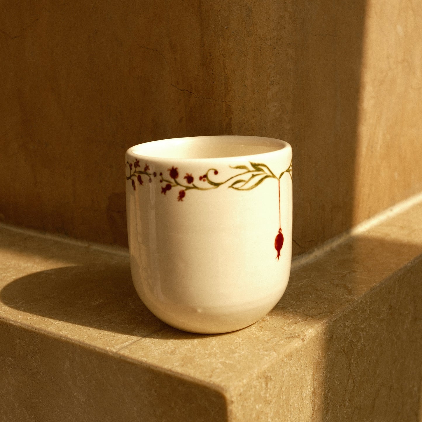 A small white ceramic cup with a delicate gold vine and red berry pattern, resting on a beige stone ledge in warm sunlight.