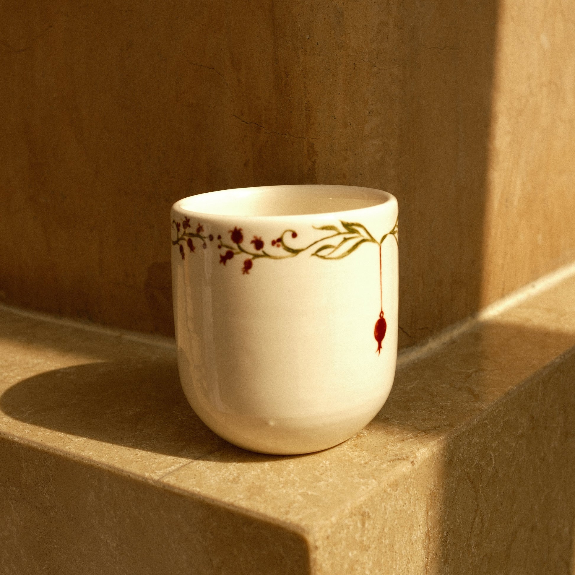 A small white ceramic cup with a delicate gold vine and red berry pattern, resting on a beige stone ledge in warm sunlight.