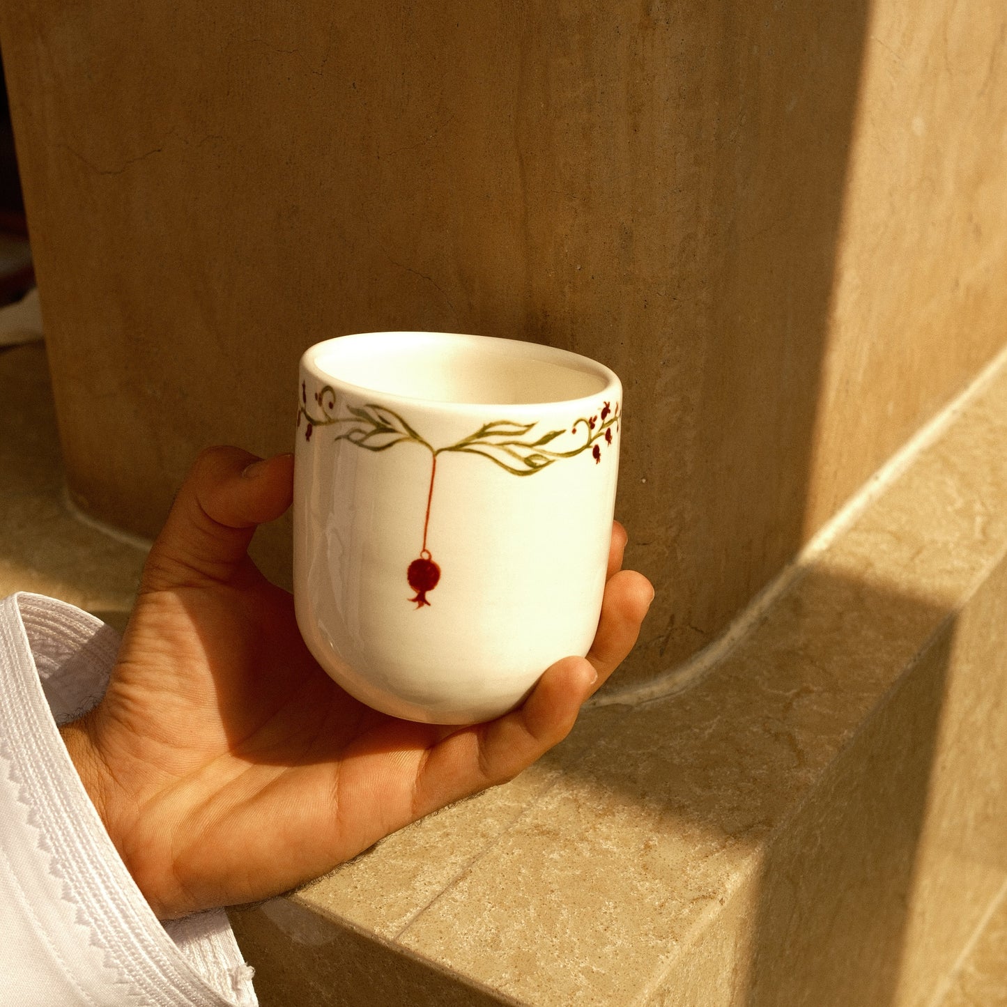 A hand holding a small white ceramic cup decorated with a gold branch pattern and a red hanging pomegranate, set against a warm beige stone background.