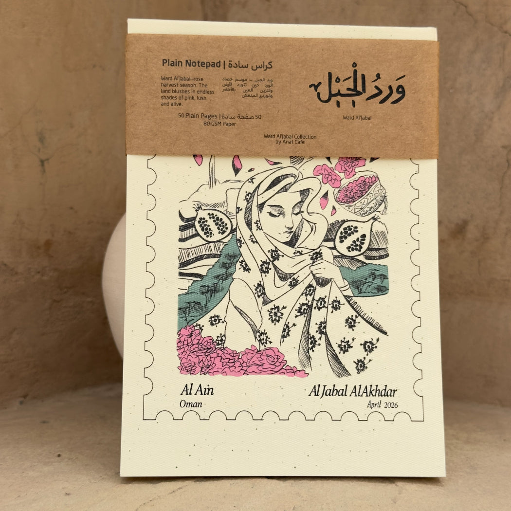 A square paper notepad with a kraft paper band across the top, photographed standing against a textured beige wall. The notepad front features a postage-stamp style border and an illustrated woman holding pomegranates in pink, green, and black linework.