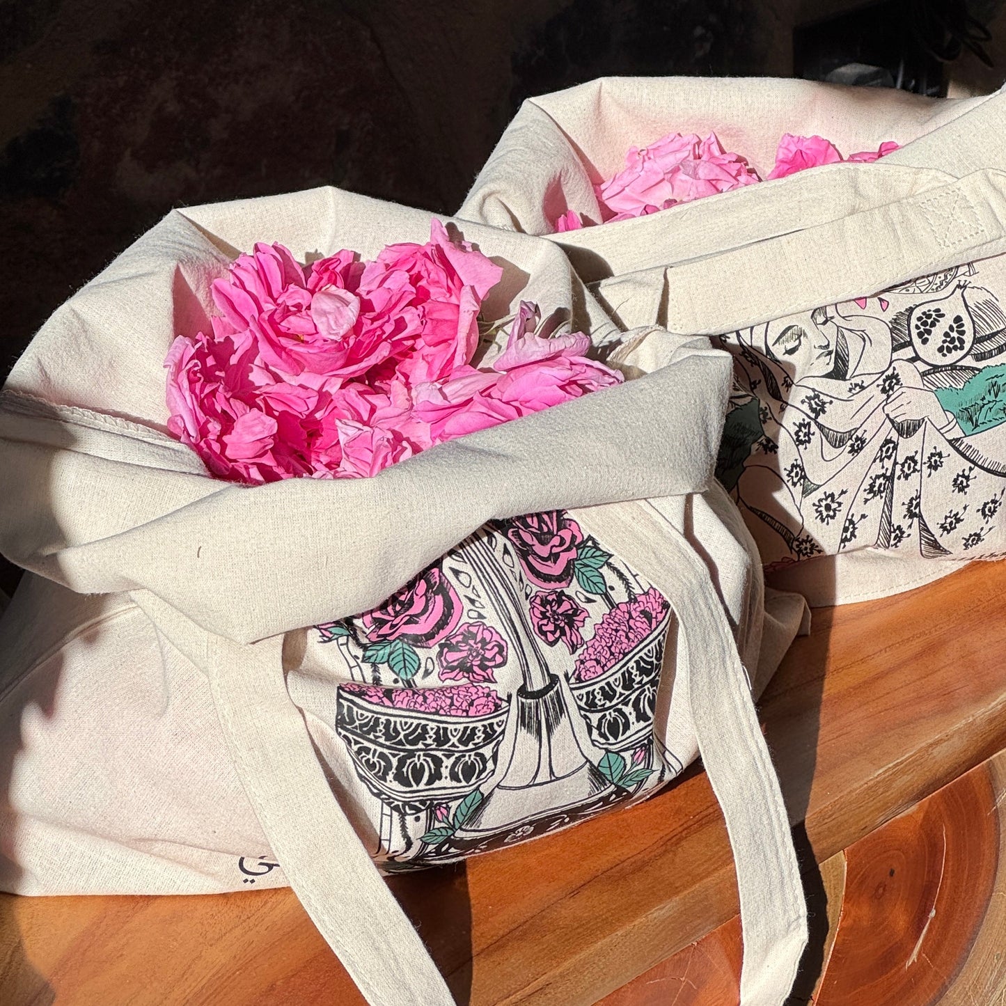 Two cream canvas tote bags filled with bright pink peony flowers, resting on a wooden surface against a dark stone wall background.